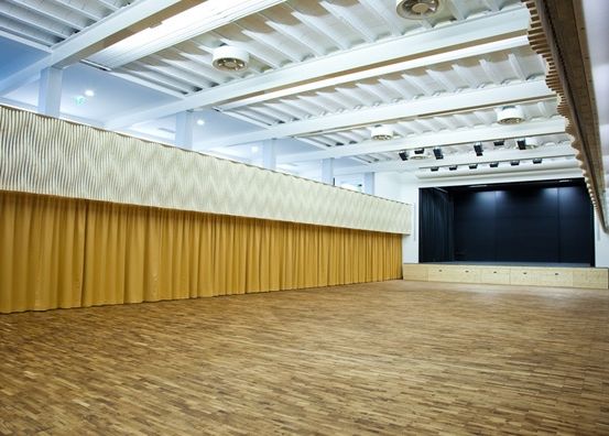 Ein leerer Raum mit Holzboden und gelben Vorhängen. Eine Bühne mit schwarzem Hintergrund ist sichtbar.