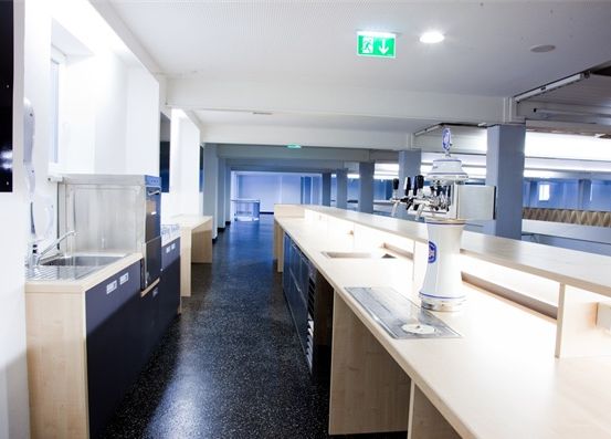 Ein leerer Küchenbereich mit weißen Arbeitsplatten, Schränken und einem Edelstahlspülbecken. Ein Bierhahn steht auf der Arbeitsplatte und ein Kühlschrank befindet sich links.