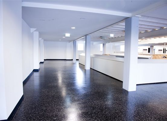 Ein leerer Raum mit weißen Wänden, schwarzem gesprenkeltem Boden und vier weißen Säulen. Ein weißer Tresen erstreckt sich entlang der rechten Seite.