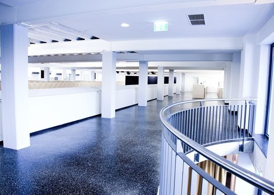 Ein leerer Büroraum mit weißen Wänden, blauem Boden und einer Metallspirale. An der Wand sind Monitore angebracht.