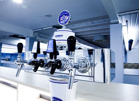 Ein Edelstahl-Getränkehahn mit zylindrischem Faß, der vier Griffe und ein rundes Emblem oben hat. Das Emblem ist blau mit weißem Text und einem roten Logo.