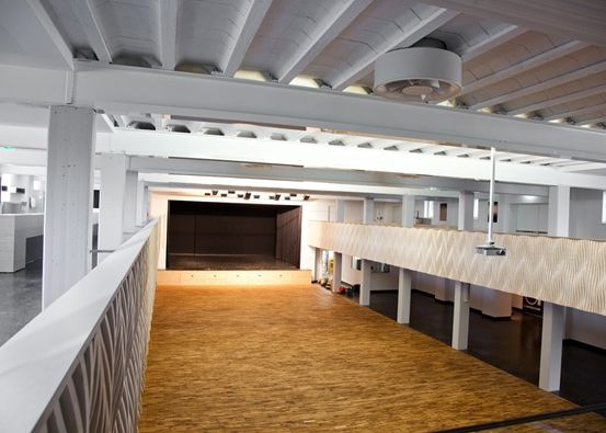 Ein leerer Saal mit Holzboden, weißen Wänden und einer Bühne mit schwarzem Vorhang. Es gibt Stützbalken an der Decke und ein an der Wand montierter Projektor.
