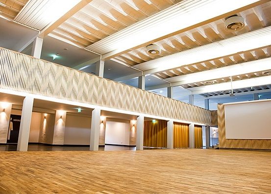 Ein leerer Saal mit Holzböden und -wänden, weißer Decke mit Leuchten, einer Galerie und einer Projektionsleinwand an der Wand.