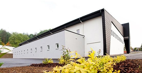 Ein modernes, weißes Gebäude mit schwarzen Akzenten, beschriftet mit 'Osterreichhalle', befindet sich in einem grünen Bereich mit Bäumen und Pflanzen. Das Gebäude hat mehrere Fenster und eine Garagentür.