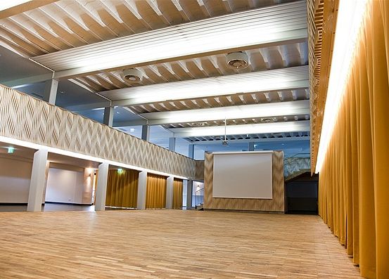 Ein leerer Saal mit Holzböden und gelben Vorhängen. Eine große Projektionsleinwand an der Wand, ohne Menschen in der Nähe.