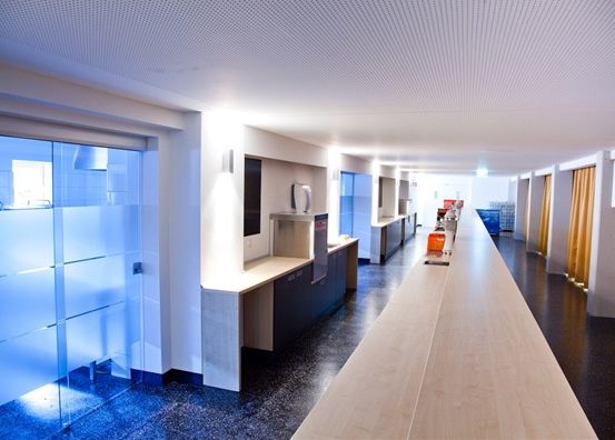 Ein leerer Innenraum eines modernen Büroraums mit einem langen Holztresen, Glaswänden, weißen Decken und beleuchteten Räumen.