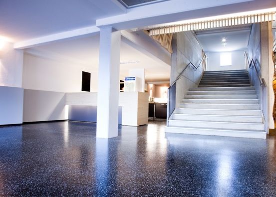 Ein leerer Raum mit weißen Wänden, einer Treppe und einem grau gesprenkelten Boden. Der Raum verfügt über eine Decke mit Lichtern und einen Monitor an der Wand.