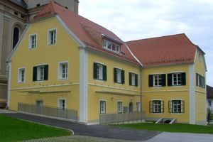 Ein großes gelbes Gebäude mit rotem Dach und grünen Fensterläden steht neben einer Kirche. Das Gebäude hat einen Rasen vor dem Haus und einen Zaun.