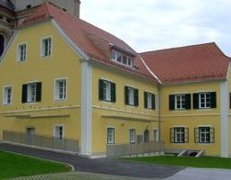 Ein großes gelbes Gebäude mit rotem Dach und grünen Fensterläden steht neben einer Kirche. Das Gebäude hat einen Rasen vor dem Haus und einen Zaun.