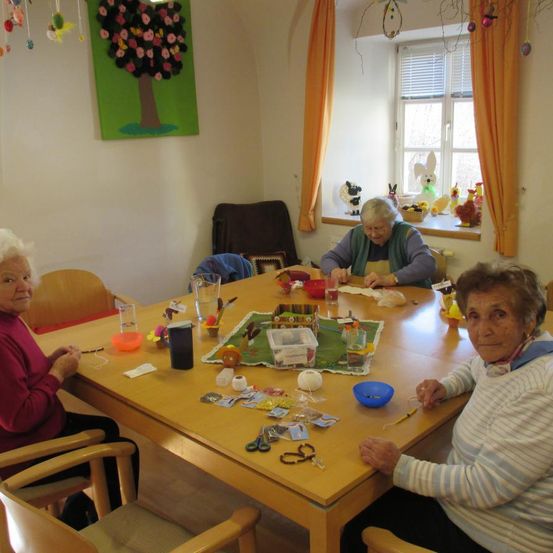 Drei ältere Frauen sitzen um einen Tisch herum und beschäftigen sich mit einer Aktivität. Sie sind von verschiedenen Gegenständen wie Scheren, Schüsseln und kleinen Spielzeugen umgeben. Ein Baumbild und ein Fenster mit Osterdekorationen sind im Hintergrund zu sehen.