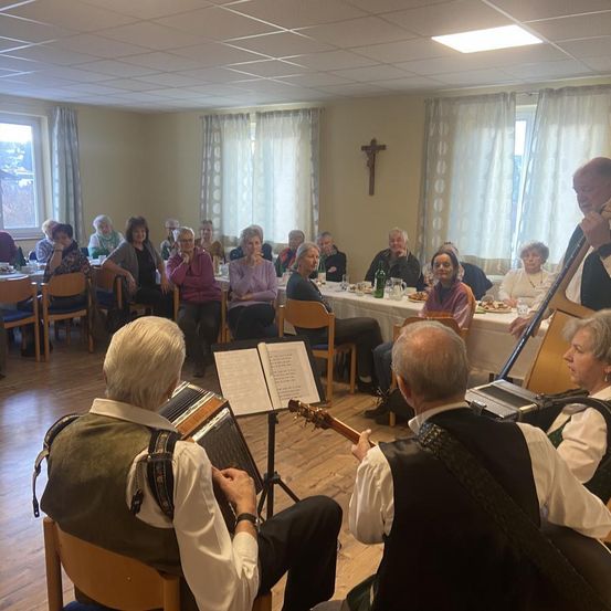 Eine Gruppe von Menschen sitzt um einen Tisch in einem Speisesaal herum, einige spielen Instrumente. Ein Kreuz an der Wand und ein Fenster mit Vorhängen.