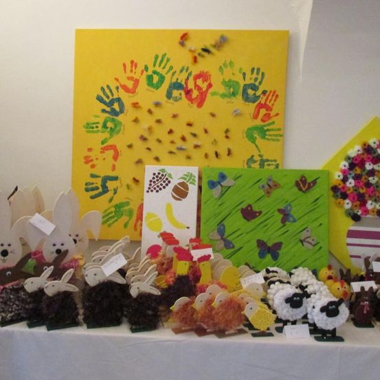 Ein Tisch zeigt handgefertigte Tierfiguren, darunter Kaninchen, Hühner und Schafe, mit bunten Postern im Hintergrund. Die Wand zeigt ein großes gelbes Poster mit Handabdrücken und ein kleineres grünes Poster mit Schmetterlingen.