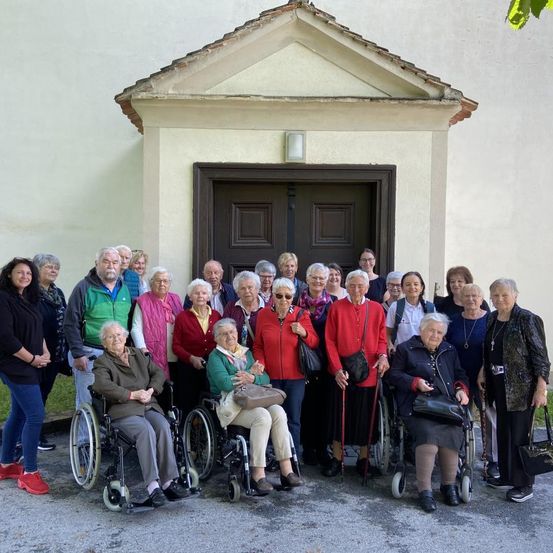 Eine Gruppe älterer Menschen, einige in Rollstühlen, steht vor einem Gebäude mit einer Frau, die eine Handtasche hält.