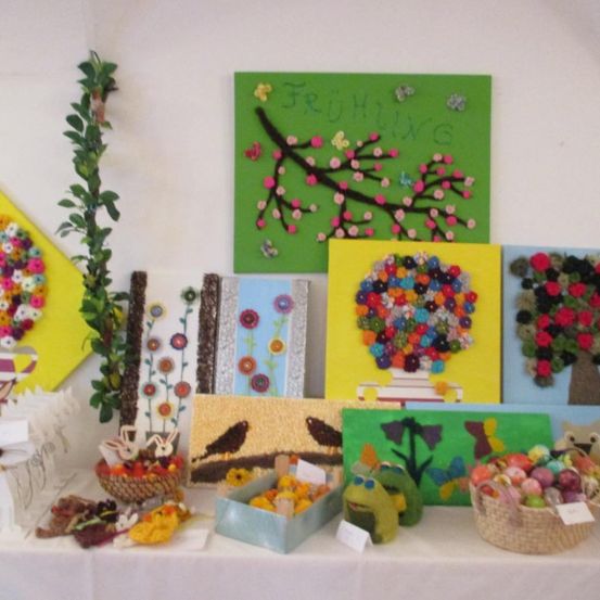 Ein Tisch zeigt eine Vielzahl von Kunstwerken, darunter gerahmte Gemälde, eine Blumenarrangement und dekorative Gegenstände. Die Gemälde zeigen verschiedene Designs, wie einen Baum, Blumen und ein Frühlingsthema. Es gibt auch Körbe gefüllt mit Gegenständen und eine Froschfigur.