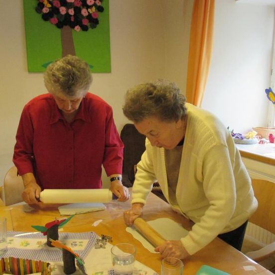 Zwei ältere Frauen arbeiten gemeinsam in einer Küche. Eine Frau benutzt einen Nudelholz auf einem Teig, während die andere an einem anderen Stück Teig arbeitet. Auf dem Tisch stehen verschiedene Gegenstände wie ein Teller, ein Glas und ein farbenfrohes Handwerk.