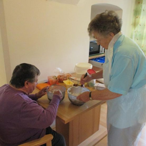 Eine ältere Frau in einem lila Hemd rührt etwas in einer Schüssel, während eine andere Frau in einem blauen Hemd daneben steht.