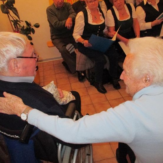 Mehrere ältere Personen sind in einem Raum versammelt. Ein Mann im Rollstuhl wird von einer älteren Frau getröstet. Andere sitzen auf dem Boden und einige halten Bücher. Ein Topf steht an der Wand.