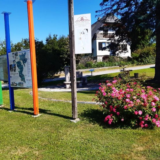 Ein grüner Bereich mit verschiedenfarbigen Stangen und einem weißen Schild mit Bild, neben einem Haus mit Balkon. Ein rosa Blumenbusch ist im Vordergrund.