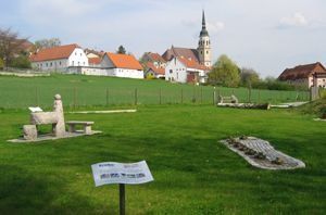 Eine ruhige ländliche Szene mit einem Kirchturm in der Ferne, einem klaren Himmel darüber und einem friedlichen grünen Feld. Es gibt mehrere Häuser, eine Bank und ein Steinmonument.