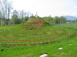 Ein kreisförmiger Garten mit einem zentralen Hügel aus Grünpflanzen, umgeben von spiralförmigen Wegen und saftigem grünem Gras. Bäume und Berge sind im Hintergrund zu sehen.