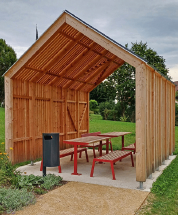 Ein hölzerner Pavillon mit einem roten Tisch und Bänken in einem Grasbereich. Der Pavillon hat ein geneigtes Dach und ist von Bäumen und Pflanzen umgeben.
