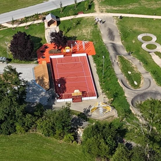 Eine Luftaufnahme eines Sportkomplexes mit einem Basketballplatz, Tennisplatz und Skatepark, umgeben von Bäumen und Gras.