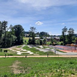 Ein Blick von oben auf einen Skatepark mit einem großen 8er-Design auf dem Boden, umgeben von grünem Gras, Bäumen und einem Tennisplatz in der Ferne.