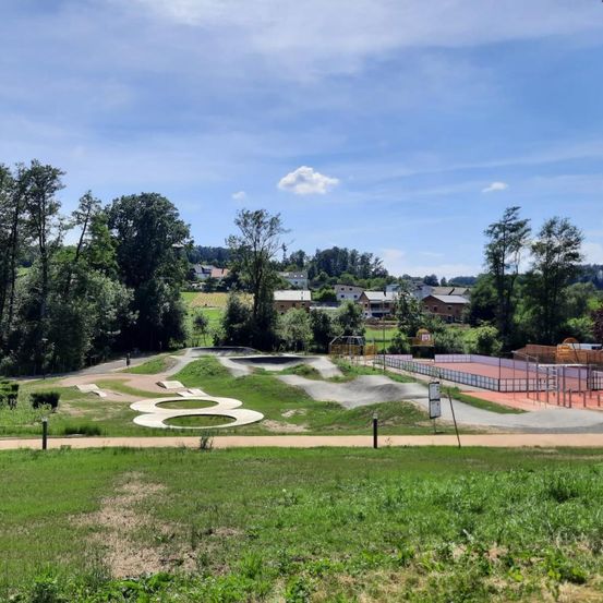 Ein Blick von oben auf einen Skatepark mit einem großen 8er-Design auf dem Boden, umgeben von grünem Gras, Bäumen und einem Tennisplatz in der Ferne.