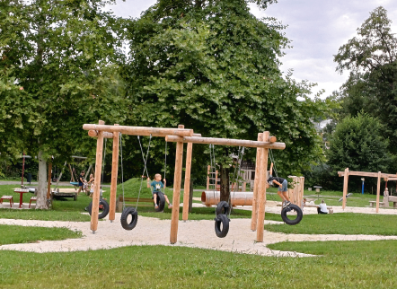 Kinder spielen auf einem hölzernen Hindernisparcours in einem Park mit Bäumen und Gras. Einige Menschen sitzen und stehen in der Nähe.