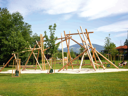 Ein Outdoor-Spielplatz mit Holzstrukturen für Kinder, umgeben von Bäumen und Gras, und einem Gebäude in der Ferne.