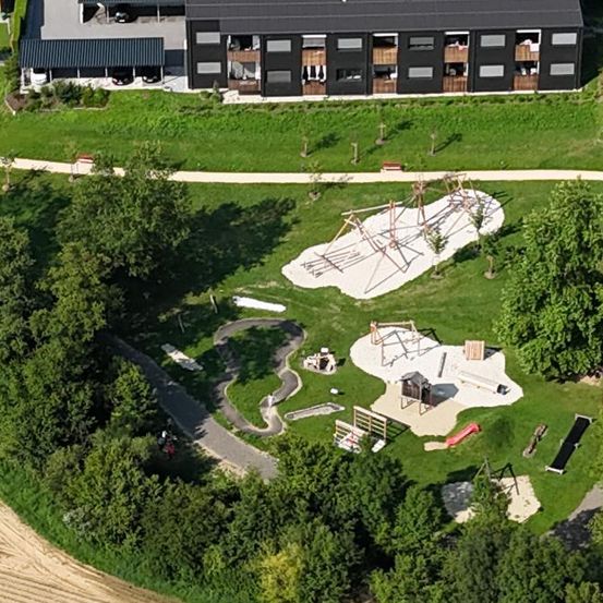 Luftaufnahme eines Wohngebäudes mit mehreren Wohnungen, einem weitläufigen Rasen und einem Spielplatz für Kinder. Das Gebiet ist von üppiger Grünfläche umgeben.