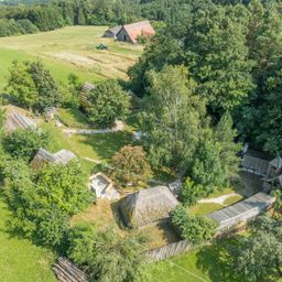 Eine Luftaufnahme einer ländlichen Landschaft mit mehreren traditionellen Häusern mit Strohdach, einem rot gedeckten Haus, einem Feld und Bäumen. Die Häuser sind von Grün umgeben.