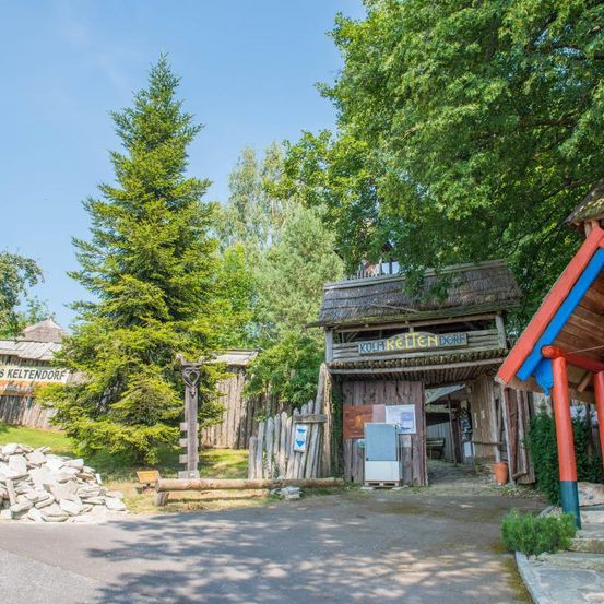 Ein kleiner Dorfeingang mit einem Schild, das Kolm Kettendorf liest, umgeben von Bäumen und einem Holzzaun. Ein Holzschild mit Herzsymbol steht links.