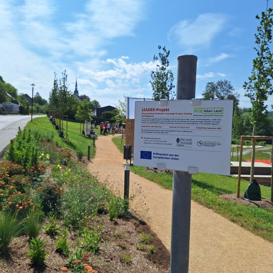Ein Schild in Englisch und Deutsch auf einem Pfosten zeigt das LEADER-Projekt für die Parkentwicklung im Sport- und Erholungsbereich.
