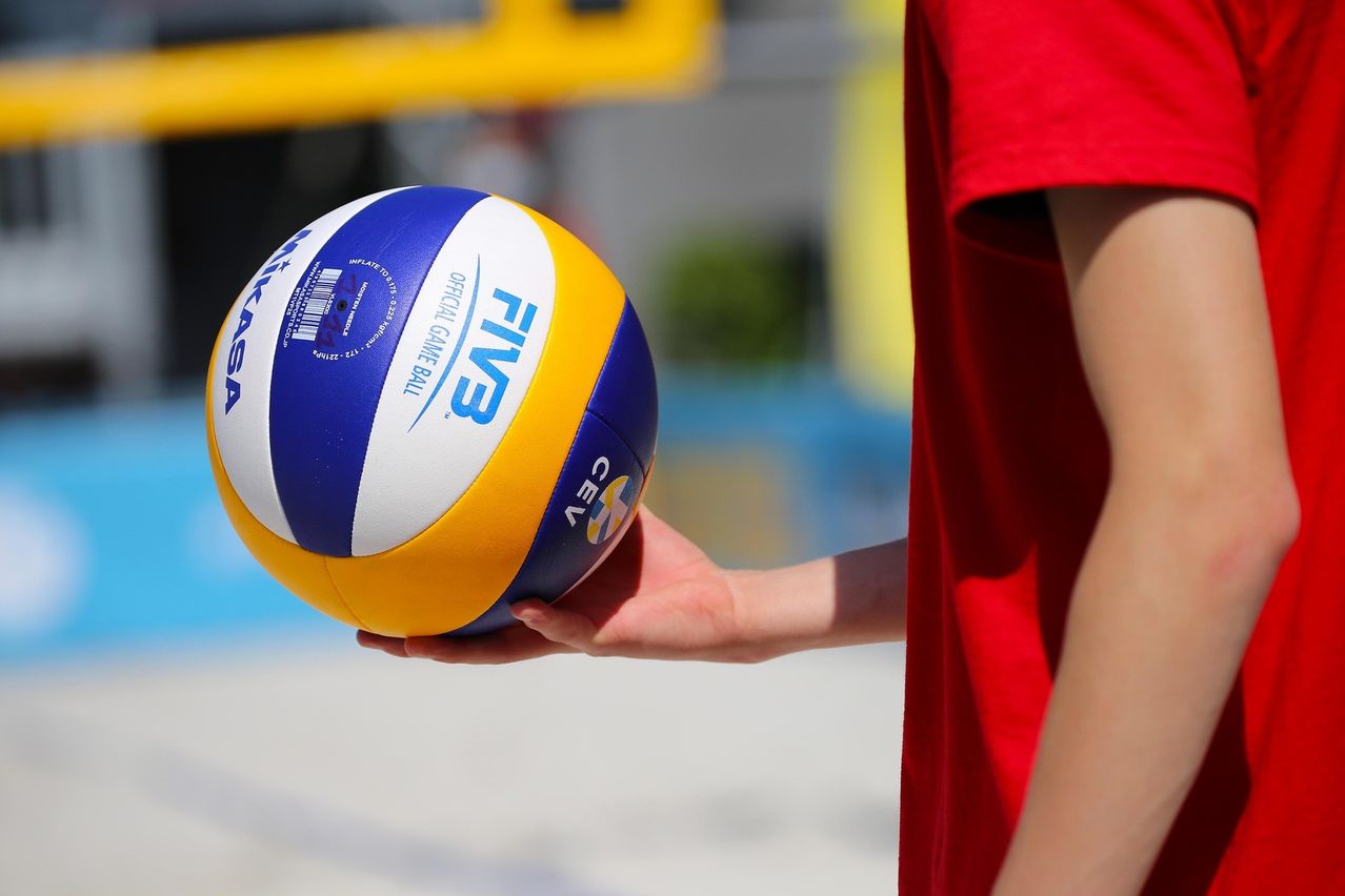 A person is holding an official FIVB beach volleyball ball. The ball is blue, yellow, and white. The background is blurry.