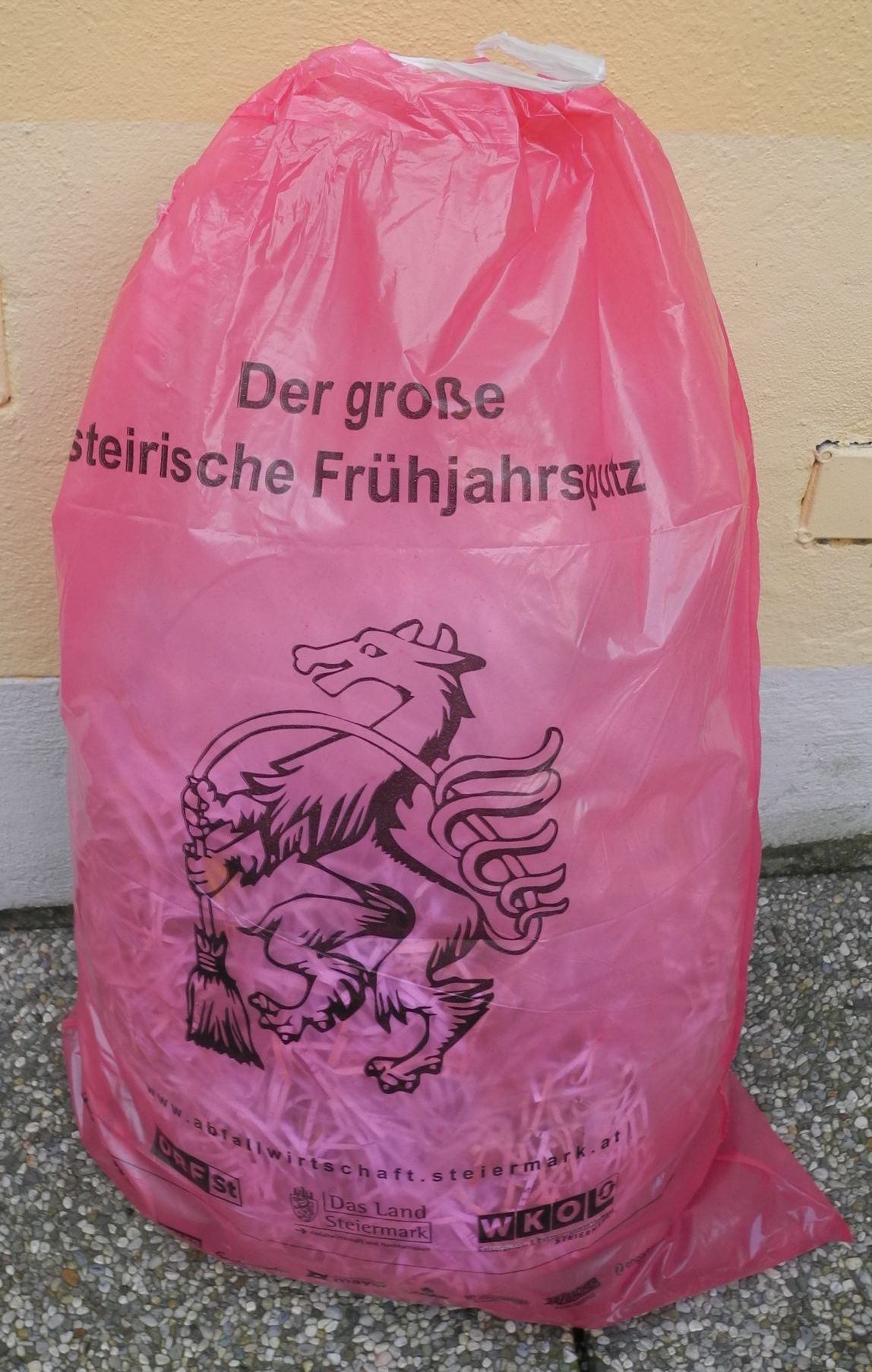 Ein rosa Plastiksack mit einem Logo eines Löwen, der einen Besen hält, beschriftet mit 'Der grosse steirische Fruhjahrsputz'. Der Sack liegt auf einer Steinoberfläche.