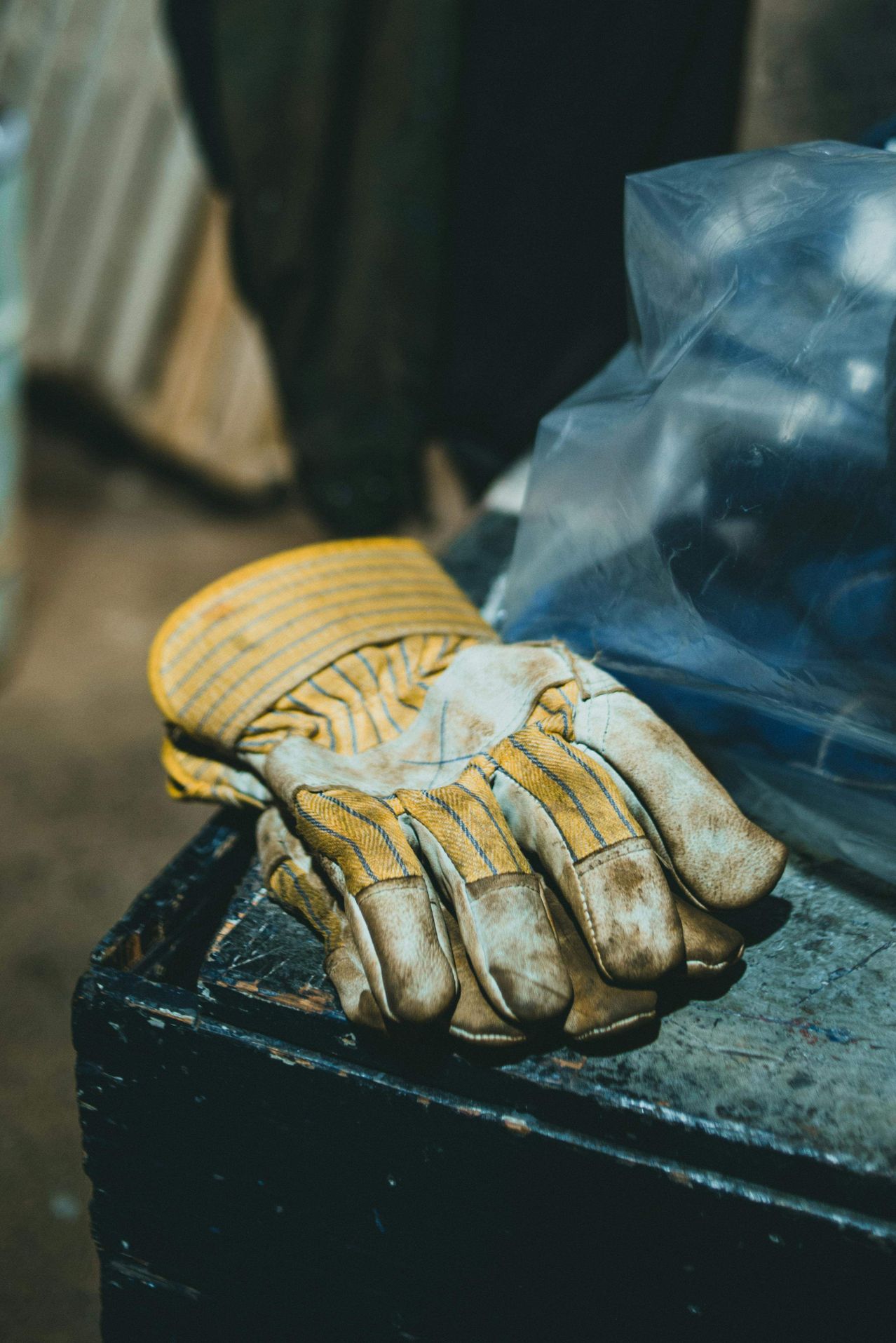 Ein Paar gelber und brauner Arbeitshandschuhe mit Schmutz und Abnutzung liegt auf einer schmutzigen, metallenen Oberfläche. Dahinter sind ein Plastikbeutel und Objekte verschwommen.