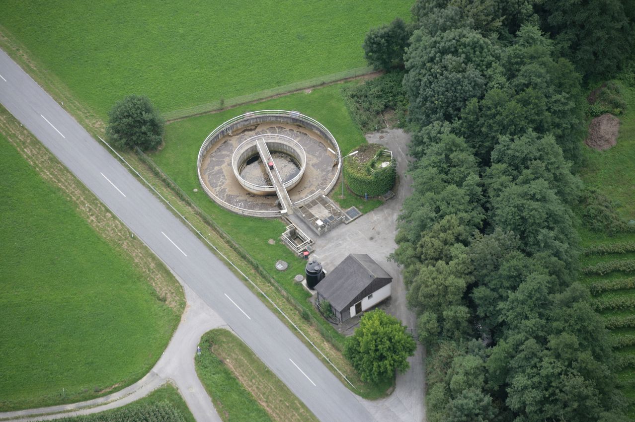 Eine Luftaufnahme einer großen kreisförmigen Kläranlage in der Nähe einer Straße, umgeben von grünen Feldern und Bäumen.
