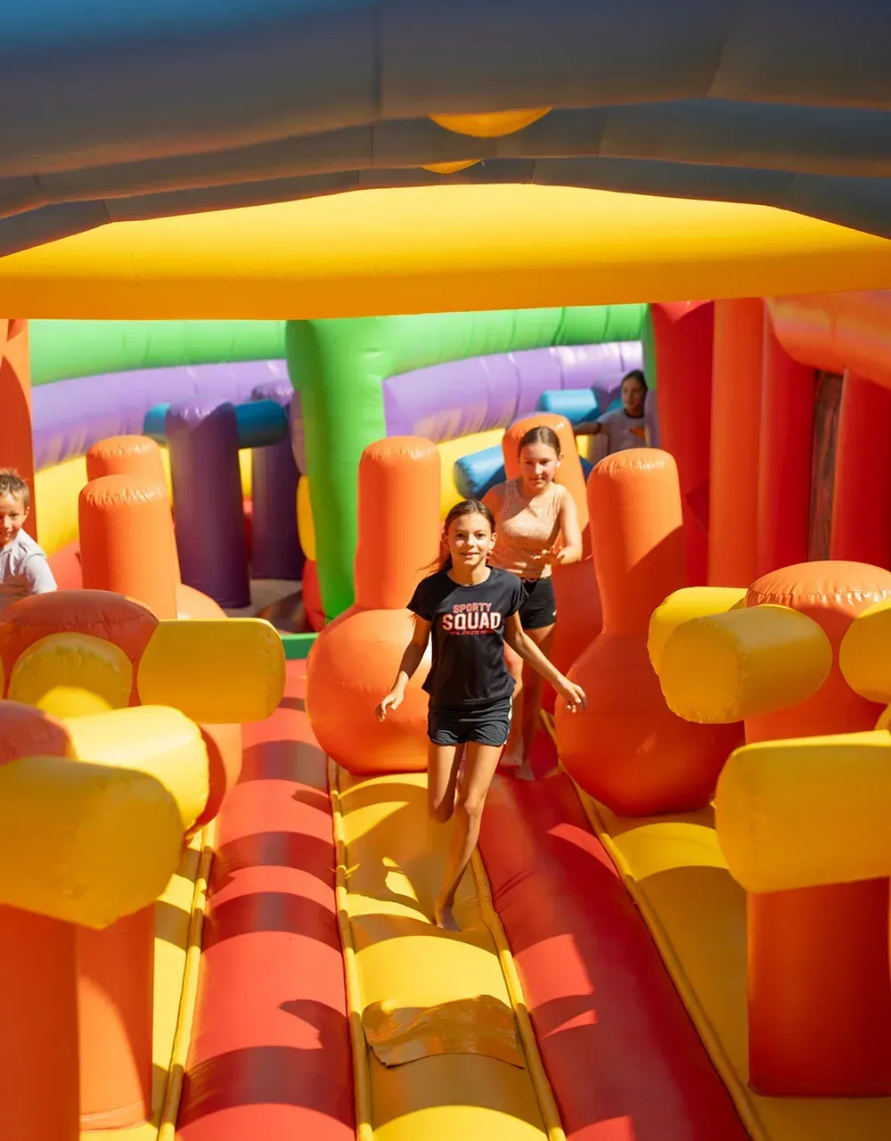 Drei Kinder spielen in einem bunten aufblasbaren Hindernisparcours. Ein Mädchen in einem schwarzen T-Shirt mit dem Wort 'SQUAD' läuft nach vorne. Hinter ihr steht ein weiteres Mädchen und beobachtet. Im Hintergrund beobachtet eine Frau.