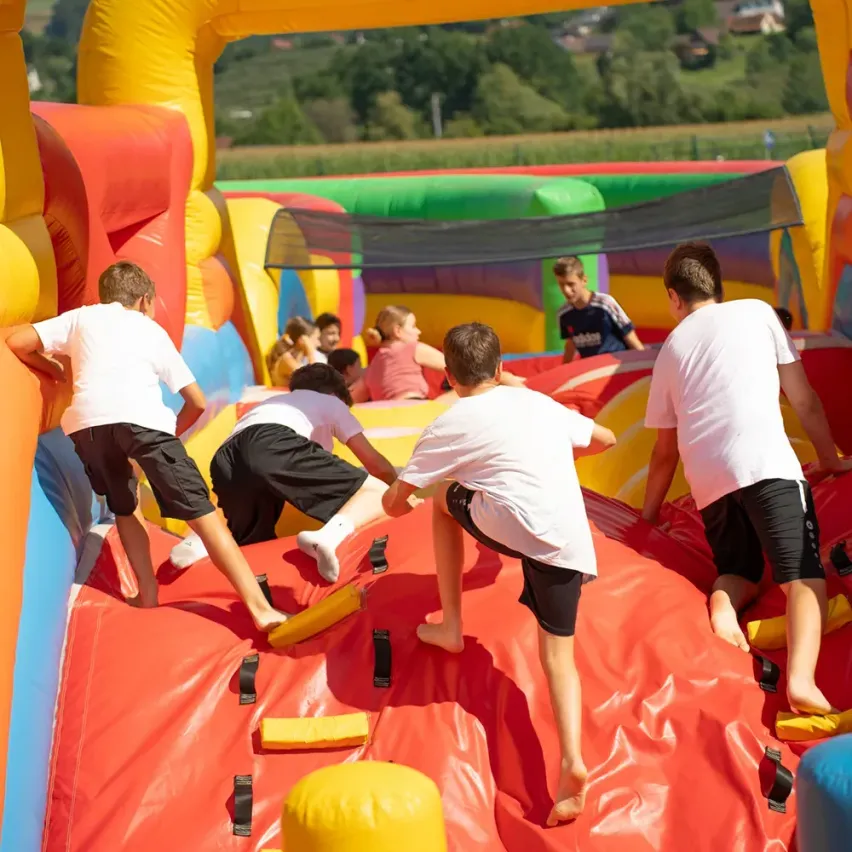 Kinder in weißen T-Shirts und Shorts klettern auf einen bunten aufblasbaren Rutschen, während andere im Hintergrund zusehen.