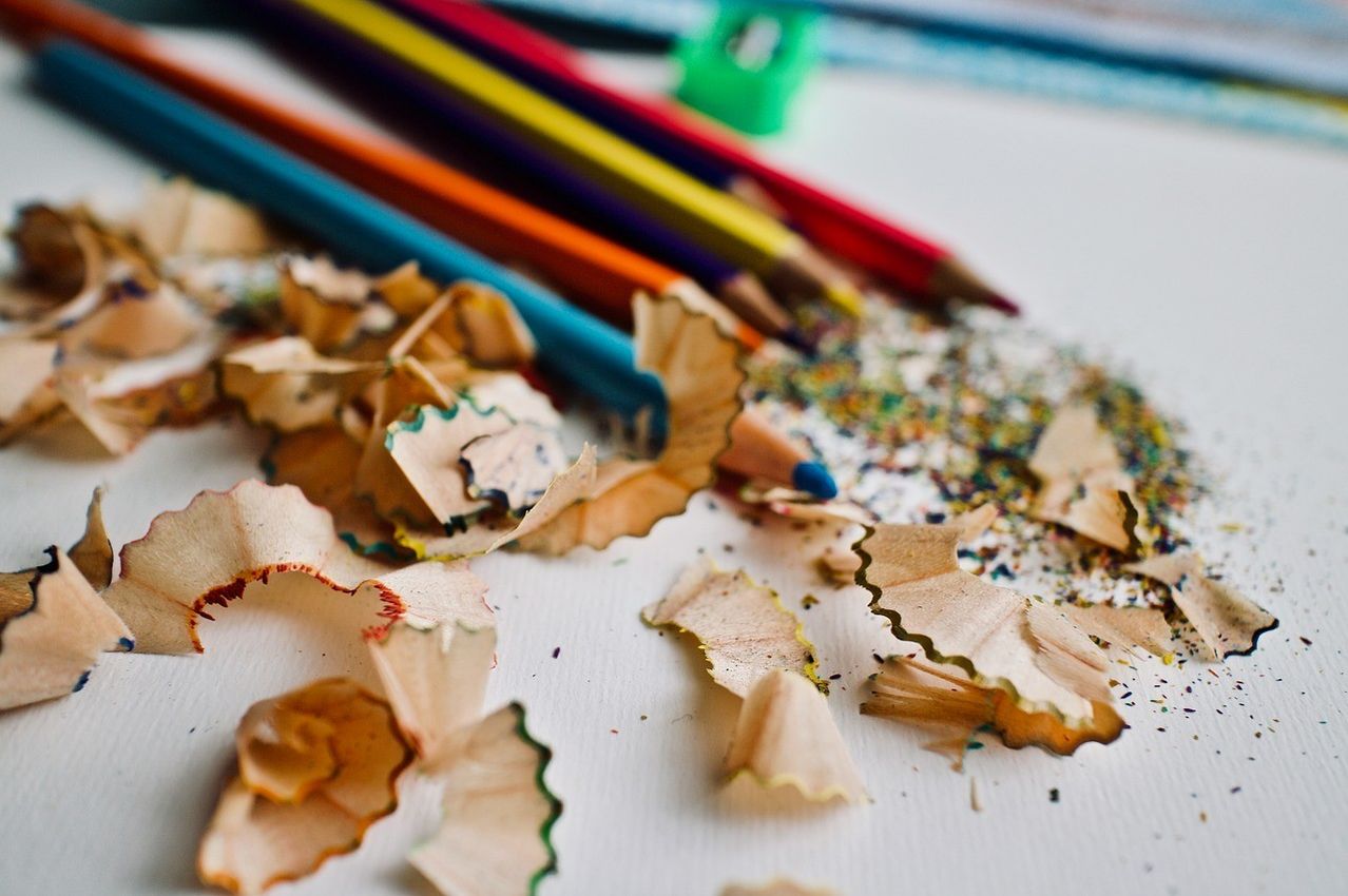 A close-up view of a white surface with various colored pencils and a pencil sharpener, surrounded by pencil shavings and small colored particles.