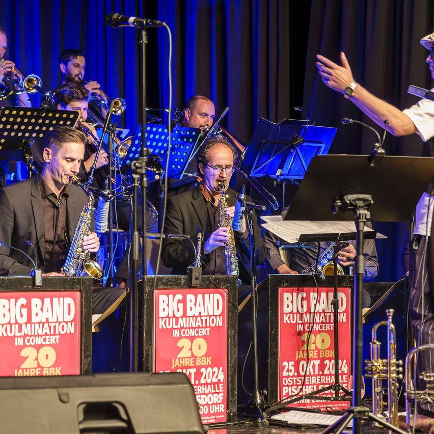 A big band concert with musicians playing saxophones and trumpets on stage, with a conductor and music stands in the background.
