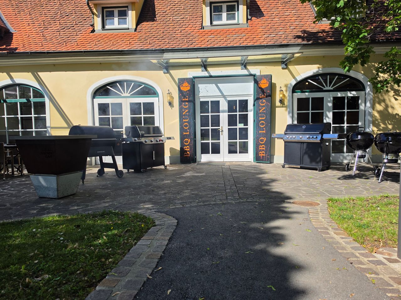 Ein gelbes Haus mit einem roten Dach und weißen Fenstern hat zwei BBQ-Grills davor. Es gibt Schilder, die BBQ Lounge sagen. Das Gebäude hat bogenförmige Fenster und eine Tür.