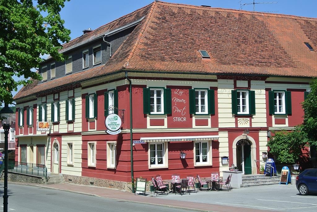 Ein rotes Gebäude mit grünen Fensterläden und einem Schild, das "Lys Neuen Post Stibor" steht. Ein Tisch und Stühle sind draußen.
