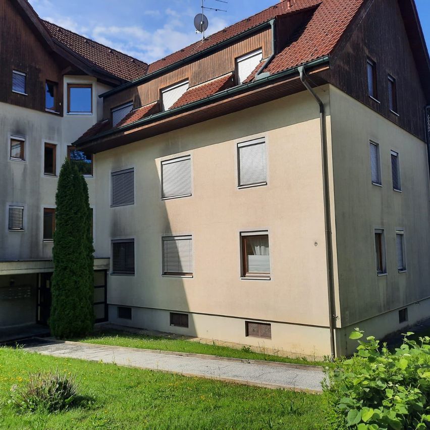 Ein dreistöckiges beiges Apartmentgebäude mit rotem Dach und braunem Rand, umgeben von Grünflächen und einer Einfahrt. Das Gebäude hat viele Fenster und einen Schornstein.