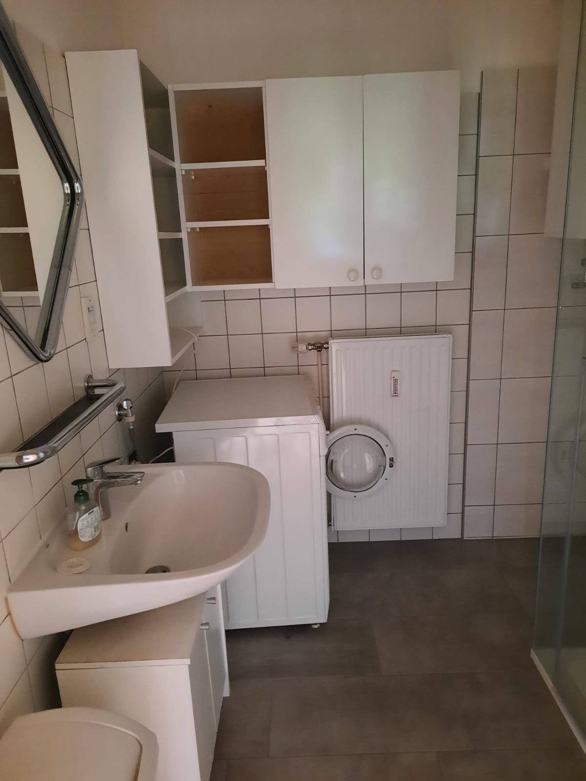 Ein Badezimmer mit weißen Fliesen, Schränken, einem Waschbecken und einer Waschmaschine. Ein Glasdusche und ein Spiegel sind vorhanden.