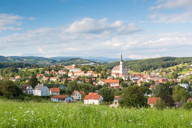 A panoramic view of a small town with red roofs, nestled among green hills under a cloudy sky.
