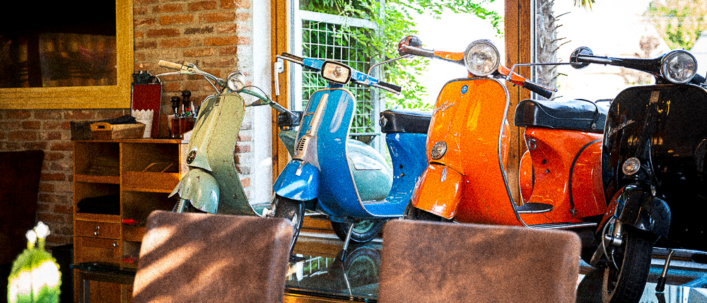 Drei Vintage-Roller sind in einem gemütlichen Raum mit einer Backsteinmauer und einem großen Fenster, das einen Garten zeigt, ausgestellt. Stühle stehen vor den Rollern.