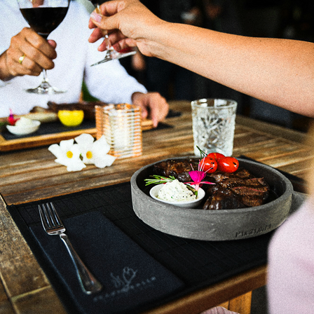 Ein Paar stößt mit Weingläsern an einem Holztisch an, auf dem sich ein Teller mit Steak befindet, das mit Kräutern und Blumen garniert ist.