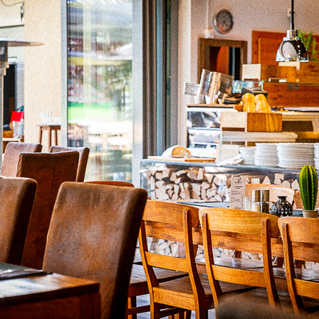 Ein Restaurant-Interieur mit Holzstühlen und -tischen, einem Kaktus in einem Topf und einer Theke mit Tellern und Brot. Eine Glaswand bietet einen Blick auf einen Außenbereich.